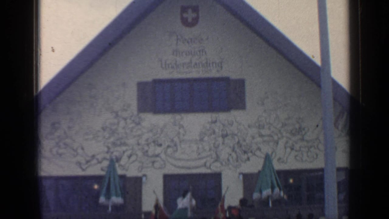 Chalet-style restaurant facade with Swiss cross, mural lettering, windows, umbrellas, and visitors below.