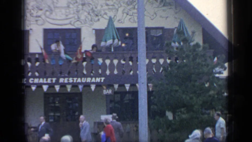 Exterior of Le Chalet Restaurant in Flushing Meadows, showing the balcony, sign, flags, green umbrellas, tree cover, and fairgoers passing below.