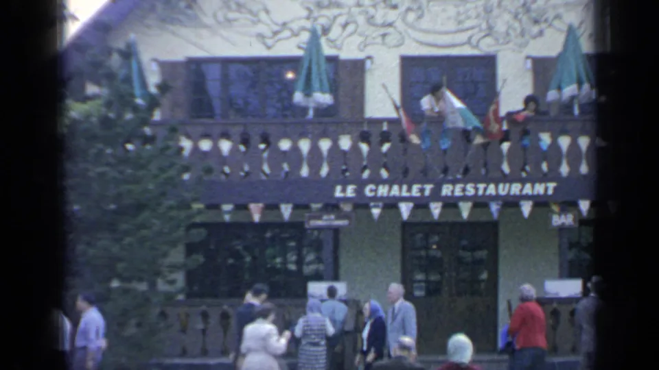 1964 Flushing Meadows visitors pass Le Chalet Restaurant, with balcony umbrellas, bunting, trees, and readable signage visible.