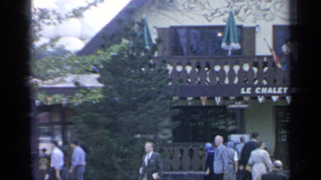 People walk past the Le Chalet Restaurant facade partly obscured by trees.