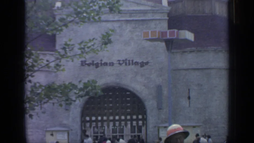 A closer 1964 view of the Belgian Village entrance shows the arched gate, wall lettering, trees, and a few visitors at the walkway edge.