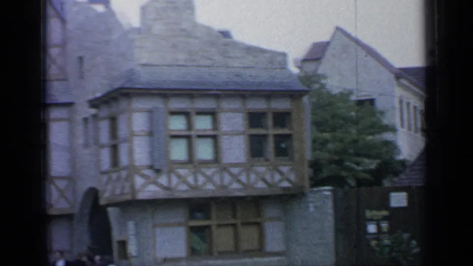 1964 Flushing Meadows home-movie frame showing a Belgian-style square with stone storefronts, half-timbered buildings, and a Belgische Wafels sign.