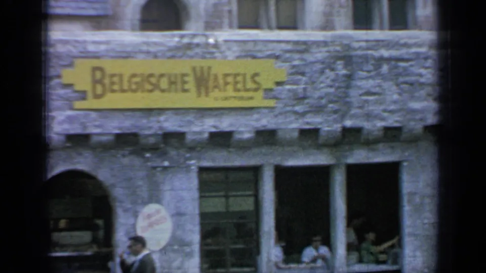1964 Flushing Meadows home-movie frame with a Belgian waffle storefront, stone wall, arched entrance, windows, and visitors gathered below.