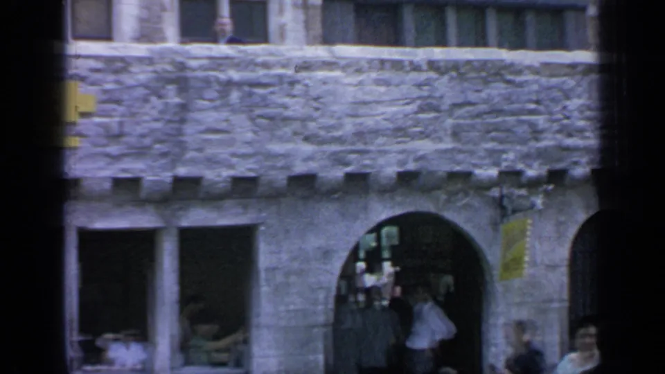 1964 Flushing Meadows frame showing a stone archway, shop windows, partial Belgische Wafels sign, and visitors moving through the Belgian-themed area.