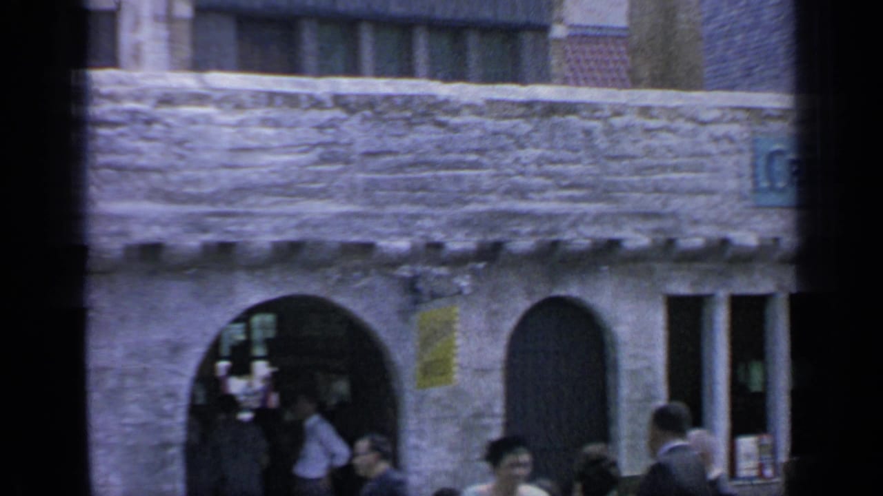 Stone storefront with arched entrances, partial shop sign, and visitors gathered below.