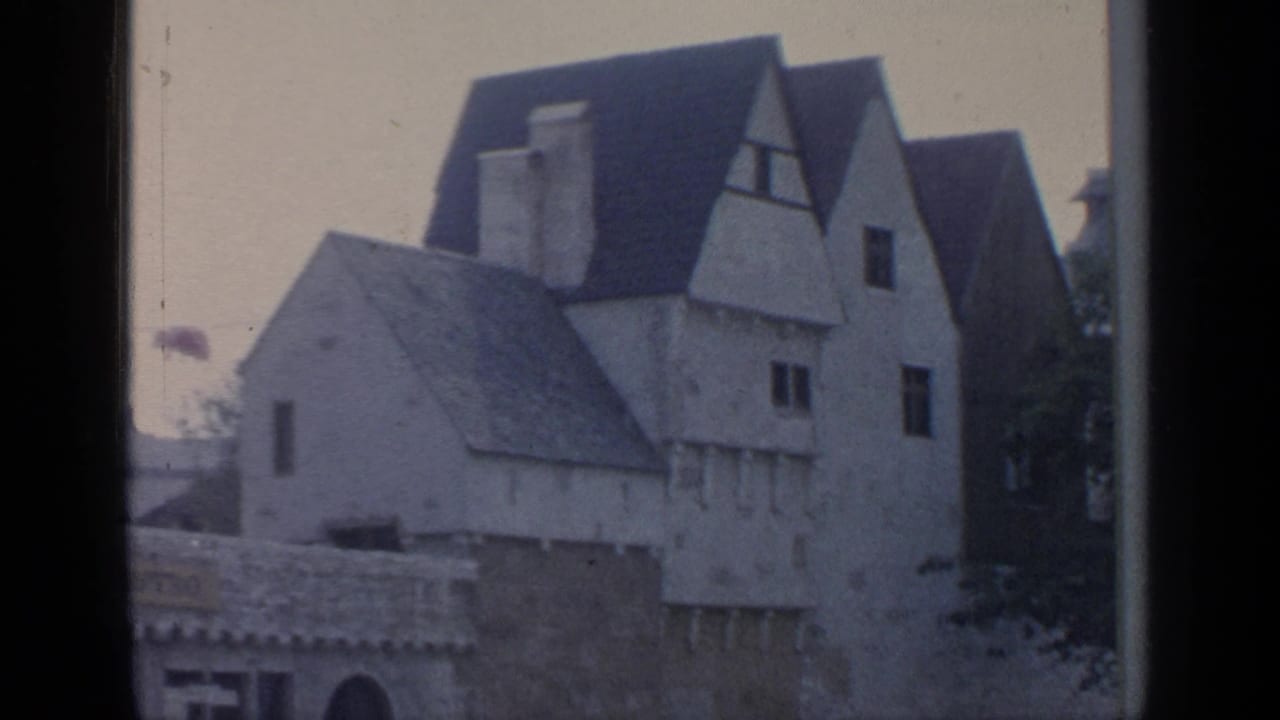 1964 8mm frame showing European-style gabled buildings and pale facades viewed through a dark window or vehicle frame.