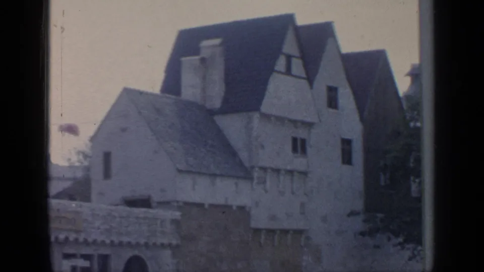 1964 8mm frame showing European-style gabled buildings and pale facades viewed through a dark window or vehicle frame.