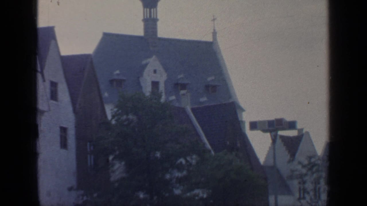 Steep-roofed building with dormers, trees and a signpost-like structure in the foreground.