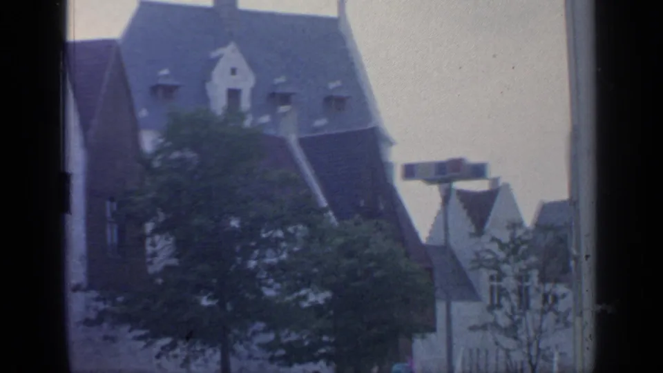 1964 8mm frame with a steep-roofed European-style building, tree canopy and smaller gables in cloudy light.