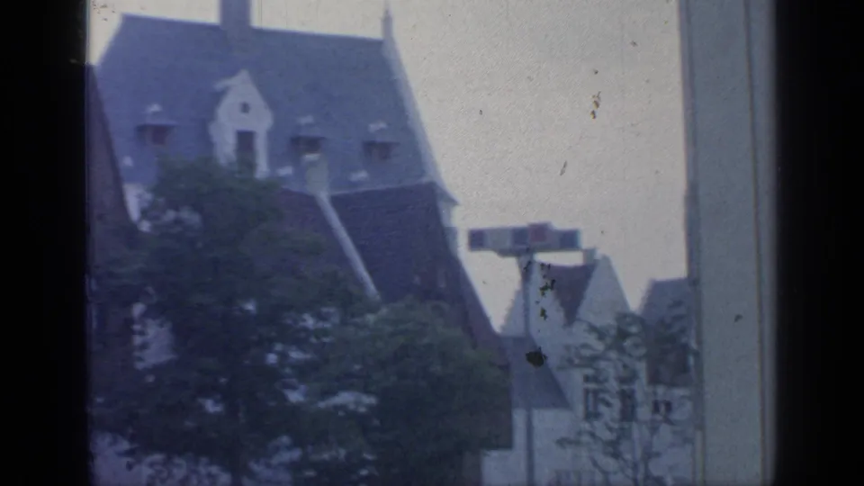1964 8mm frame showing a steep-roofed European-style building, tree branches and foreground window-frame edges.