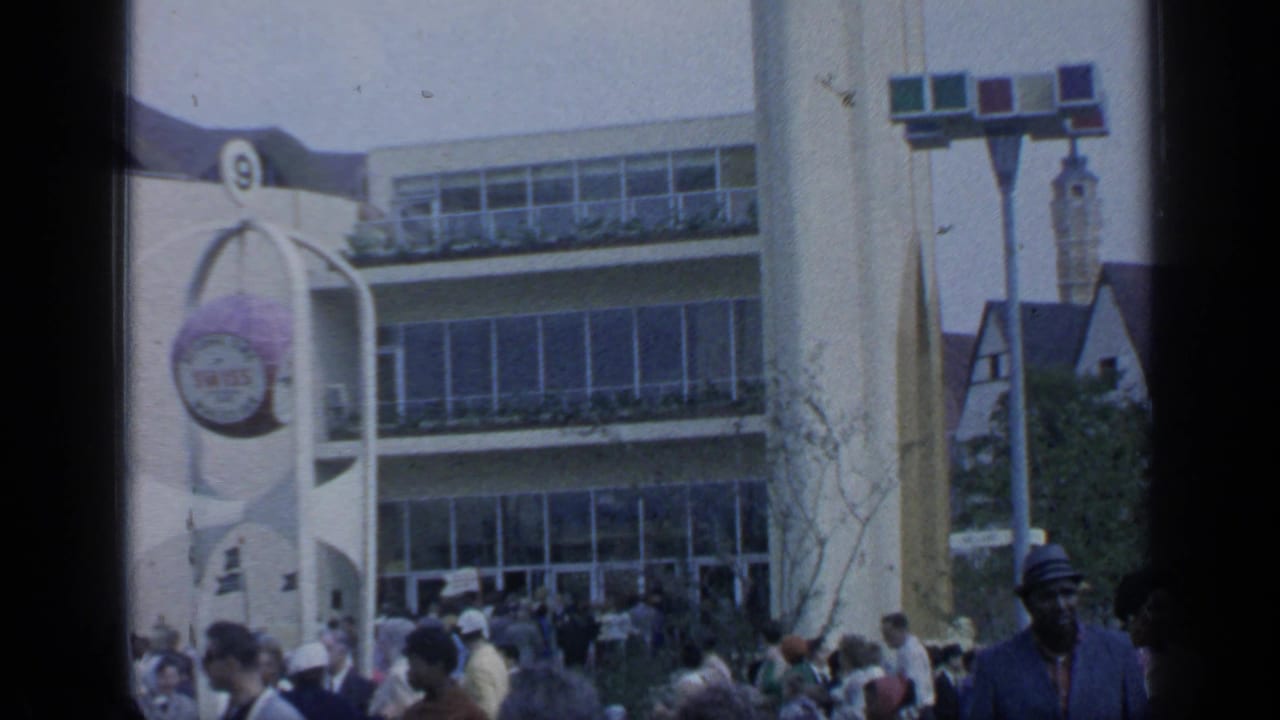 1964 Flushing Meadows frame with visitors outside a white curved fair building with a round sign and large windows.