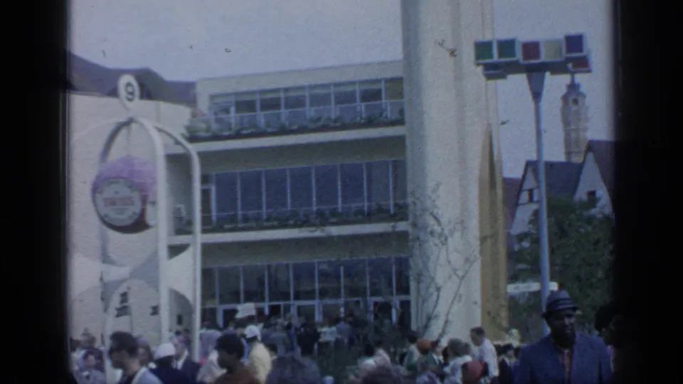 1964 Flushing Meadows frame with visitors outside a white curved fair building with a round sign and large windows.