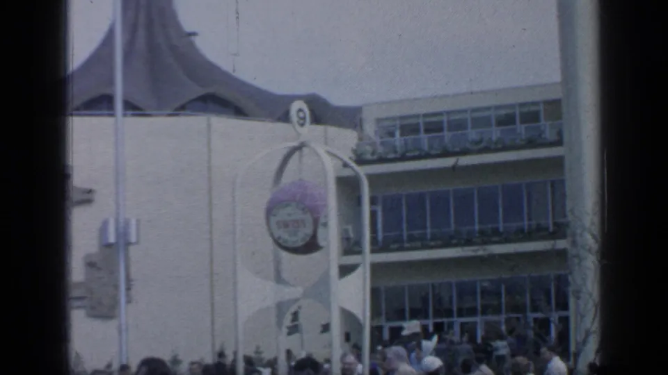 1964 Flushing Meadows frame showing a white exposition building, spherical sign, flags or poles, and visitors at the entrance.