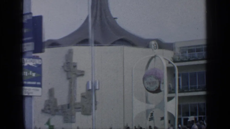 1964 Flushing Meadows frame showing curved fair architecture, a round sign, sculptural wall forms, poles, and visitors at the edge.