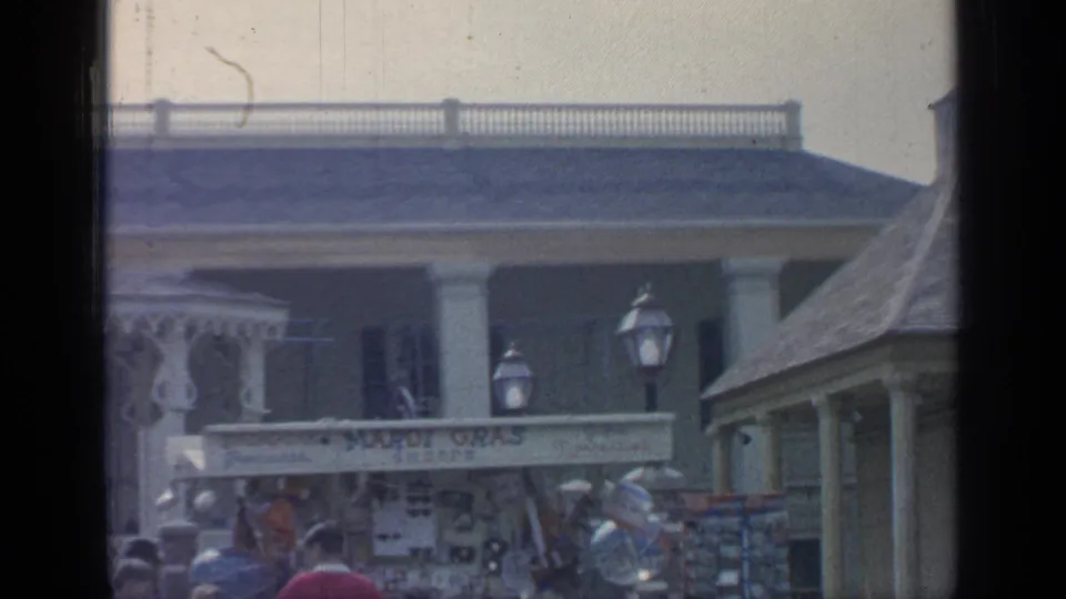 1964 Flushing Meadows frame with Mardi Gras Shoppe signage, visitors, balloons, souvenir racks, lampposts, and decorative building facades.