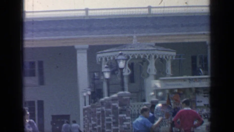 1964 Flushing Meadows pan frame showing columns, ornate gazebo trim, lampposts, brick posts, and tourists near the souvenir shop edge.