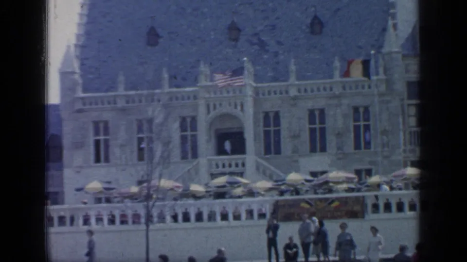 A large fairground building at Flushing Meadows appears behind terrace umbrellas, pedestrians, railings, and an American flag.