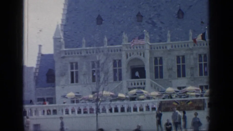 1964 Flushing Meadows home-movie frame showing a pale ornate building with flags, terrace umbrellas, and visitors below.