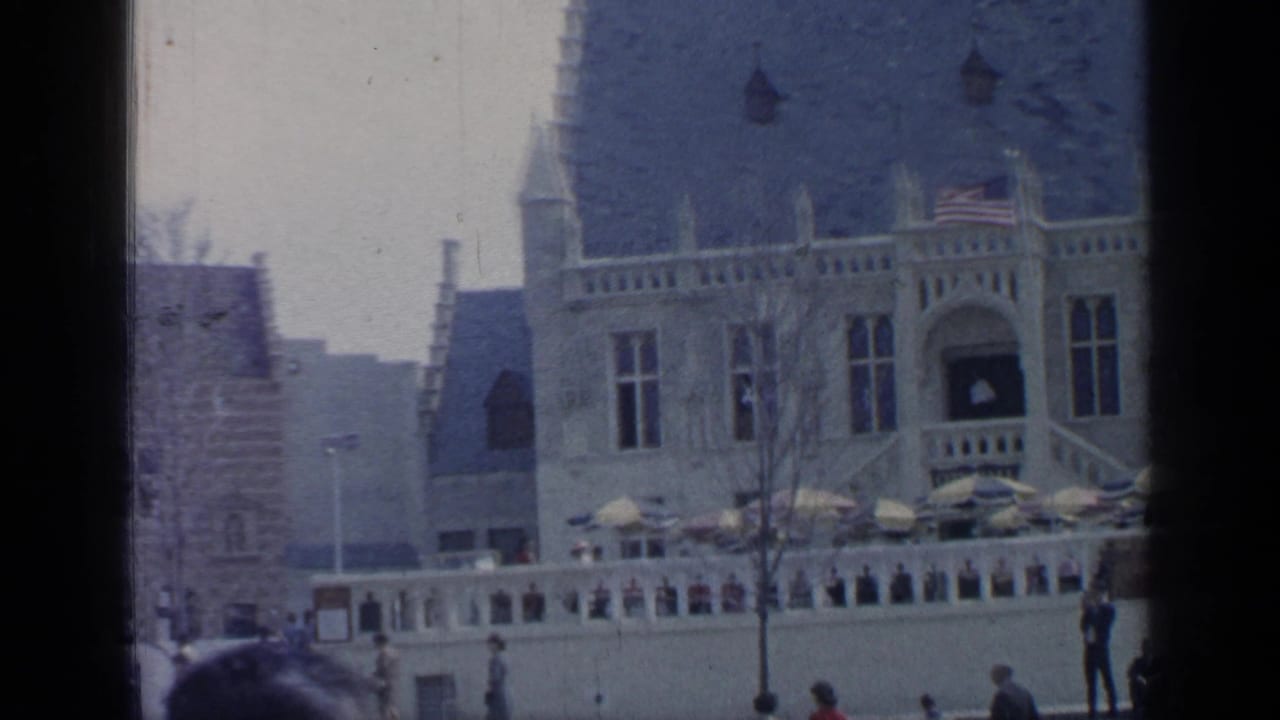 Building facade with flags, umbrellas, low wall, and people along the walkway.