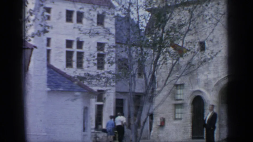 1964 Flushing Meadows home-movie frame showing visitors walking near pale stone facades, arched doors, and courtyard trees.