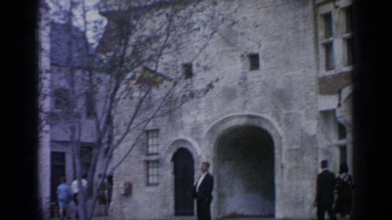 A suited man stands near an arched entrance while visitors walk in the background.