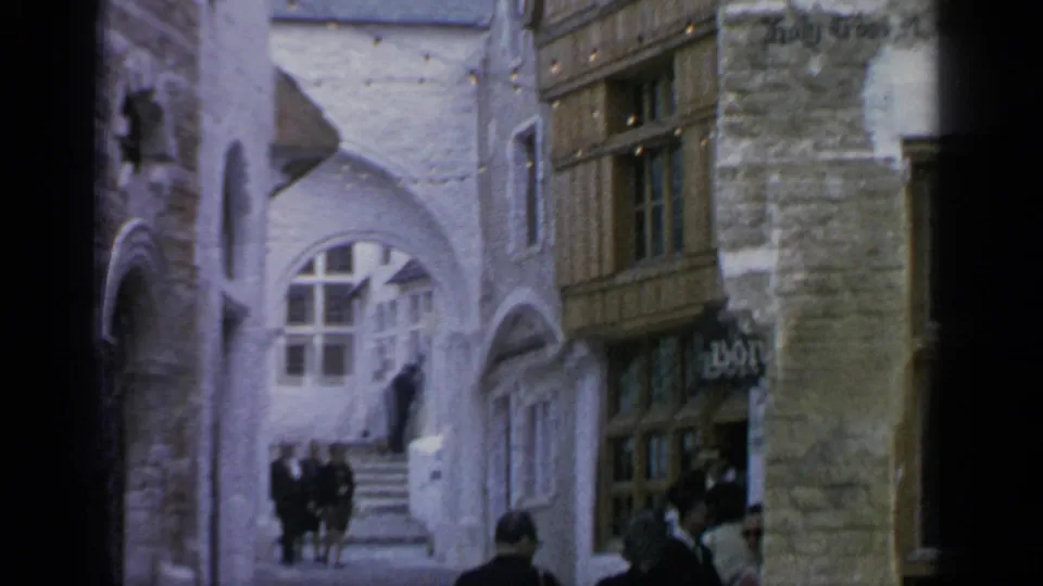 1964 home-movie frame of visitors in coats walking through a themed village passage with arches, timber facade, and stone walls.