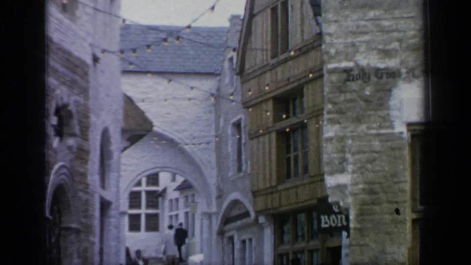 The themed World's Fair village street shows string lights, stone and timber set buildings, arched openings, and a pedestrian in the distance.
