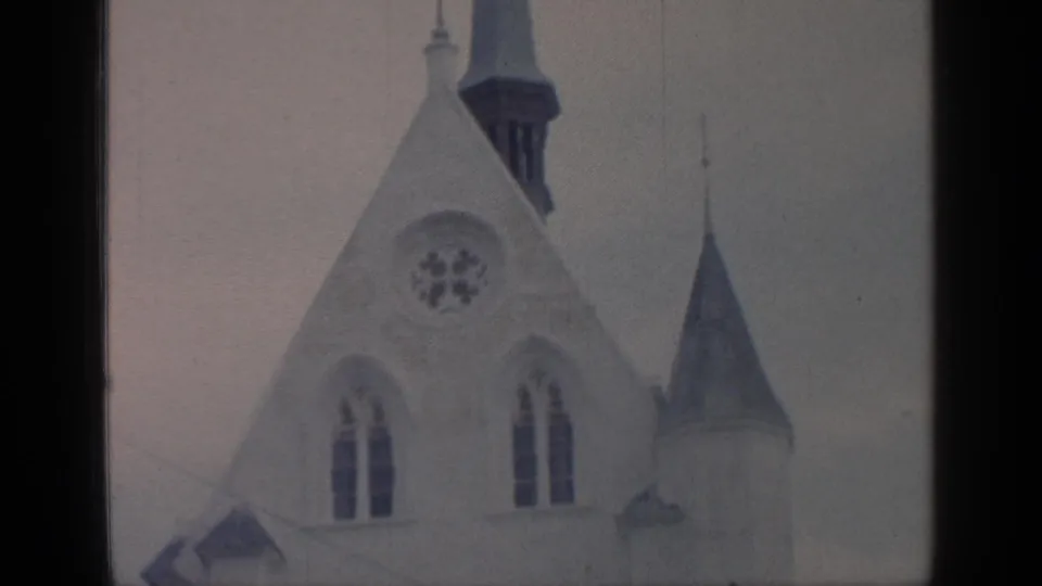 1964 Flushing Meadows frame of a pale Gothic-style church roofline, circular rose window, arched windows, spire, and cross against cloud cover.
