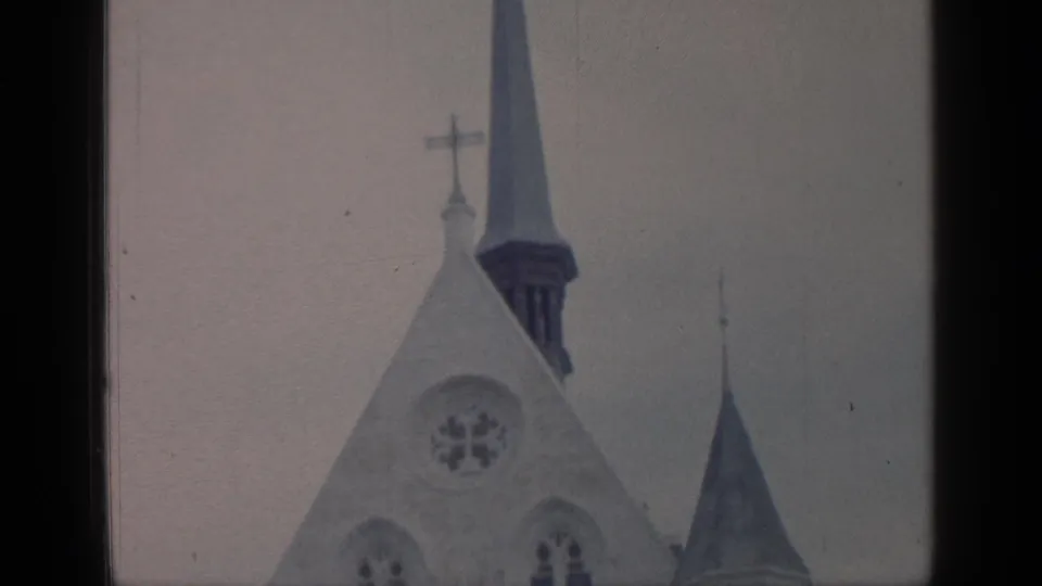 Visitor-shot 1964 Flushing Meadows frame showing a tall church spire, cross finial, circular window, and pale facade in soft 8mm color.