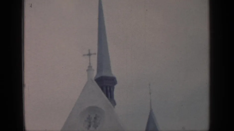 1964 Flushing Meadows home-movie frame looking up at two pointed church spires, a cross, and a pale Gothic-style facade under gray sky.