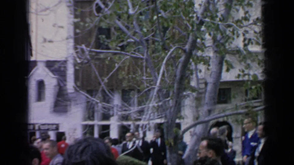 A crowd in period coats stands beneath trees near a building and tower, consistent with the 1964 Flushing Meadows home-movie setting.