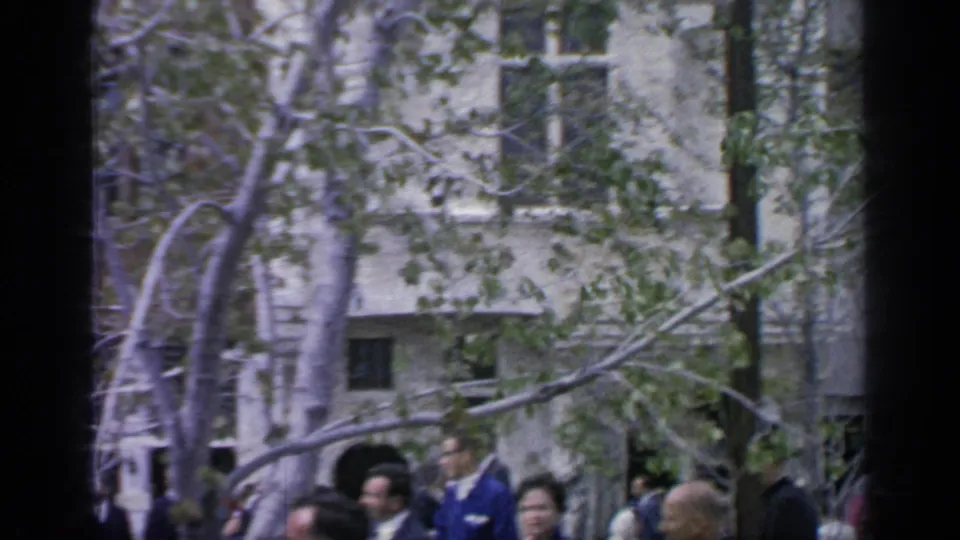 Visitors appear among trees in front of a pale building facade, with soft 8mm color and handheld framing from the Flushing Meadows reel.