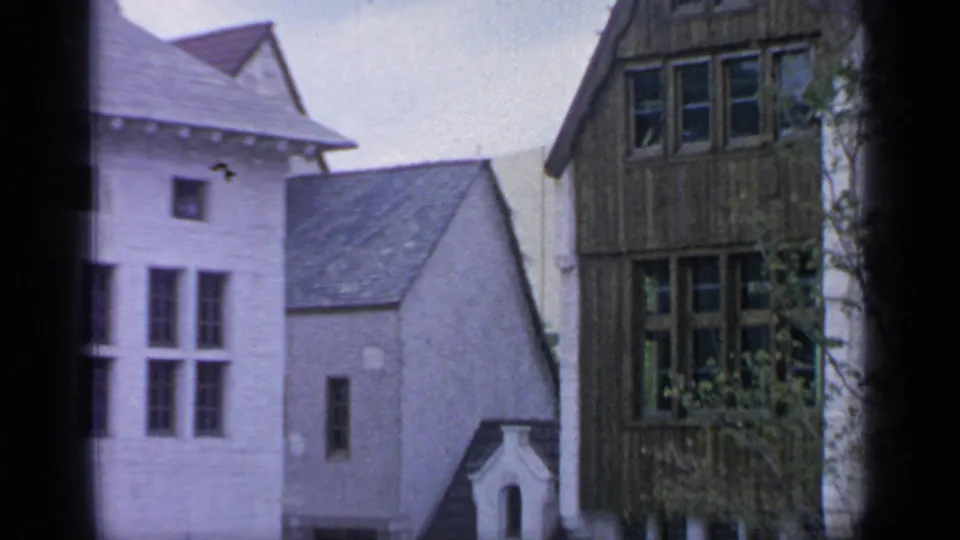 Flushing Meadows village-street view with white walls, dark timbered facade, pitched roofs, and trees in 1960s 8mm footage.