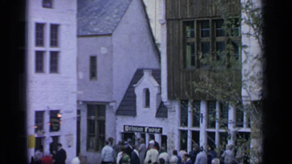 1964 Flushing Meadows crowd scene with fairgoers walking beneath village-style buildings and a Belgian Fudge shop sign.