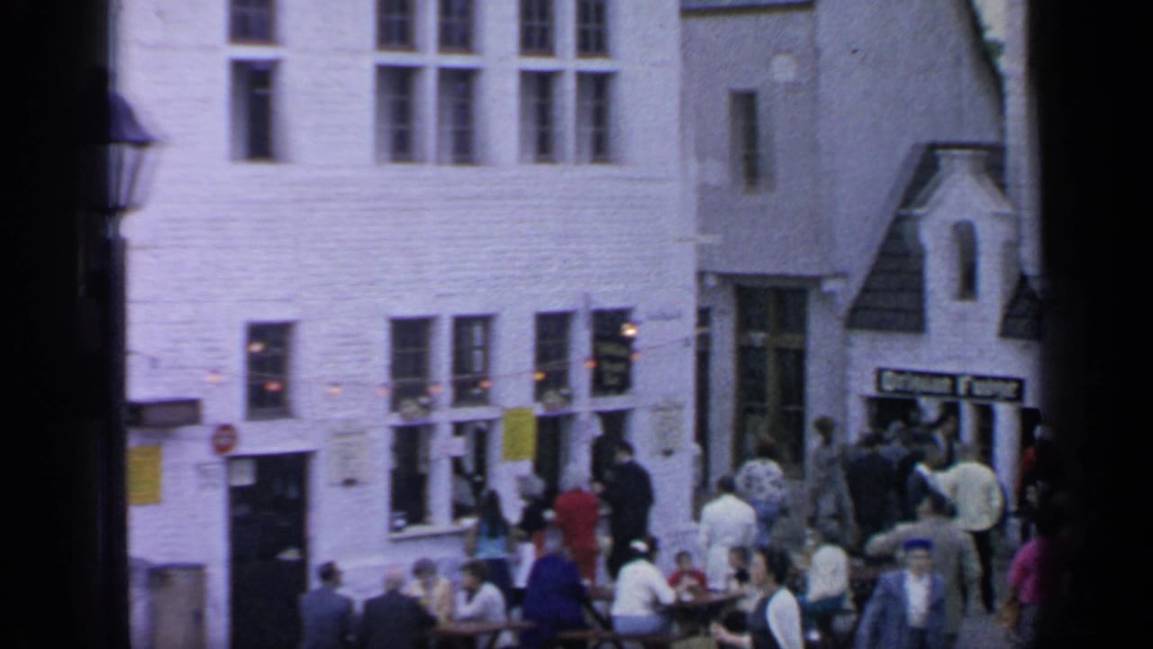 1964 Flushing Meadows street view with outdoor dining, pedestrians, white brick buildings, and small signs visible near the entrance.