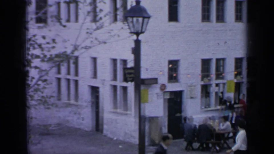 Flushing Meadows street scene with a black streetlamp, white brick building, outdoor table, and people seated or walking nearby.