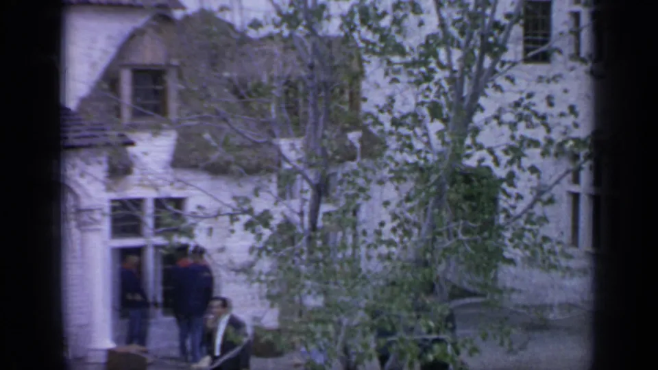 End frame from the 1964 Flushing Meadows street scene, showing a doorway, standing people, trees, windows, and white brick facades.