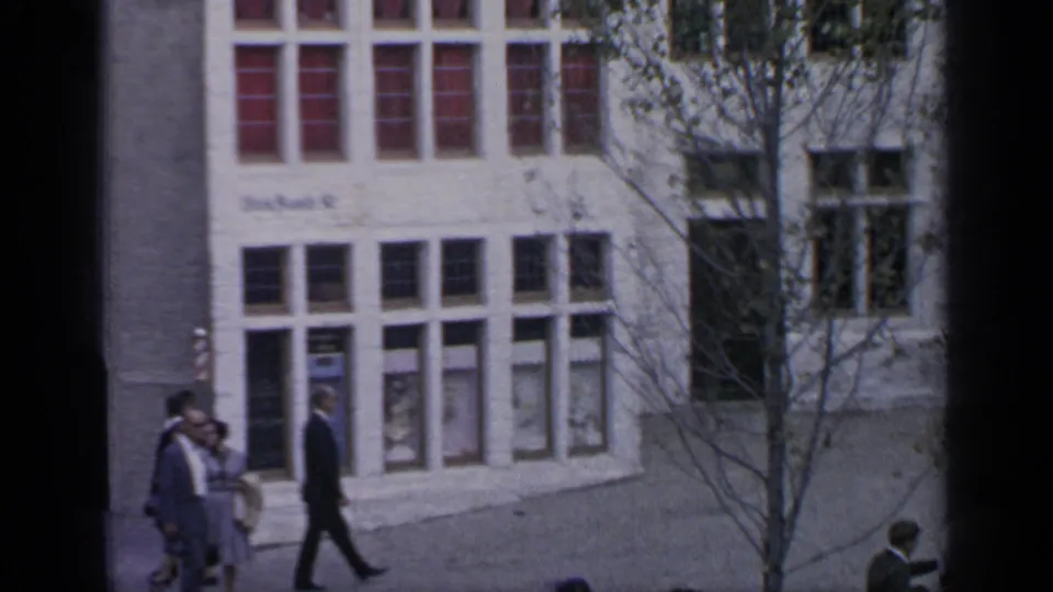 1964 Flushing Meadows street scene showing pedestrians beside a pale building with tall windows and a tree in the foreground.
