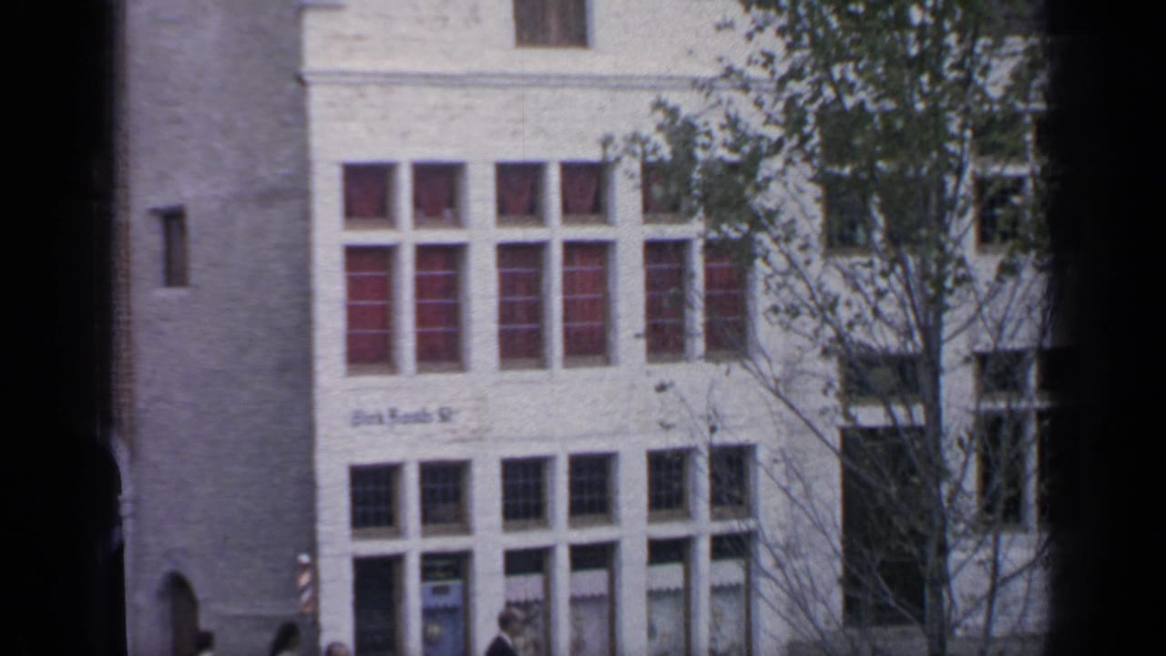 Pale building facade with red-tinted windows, trees and pedestrians near the street level.
