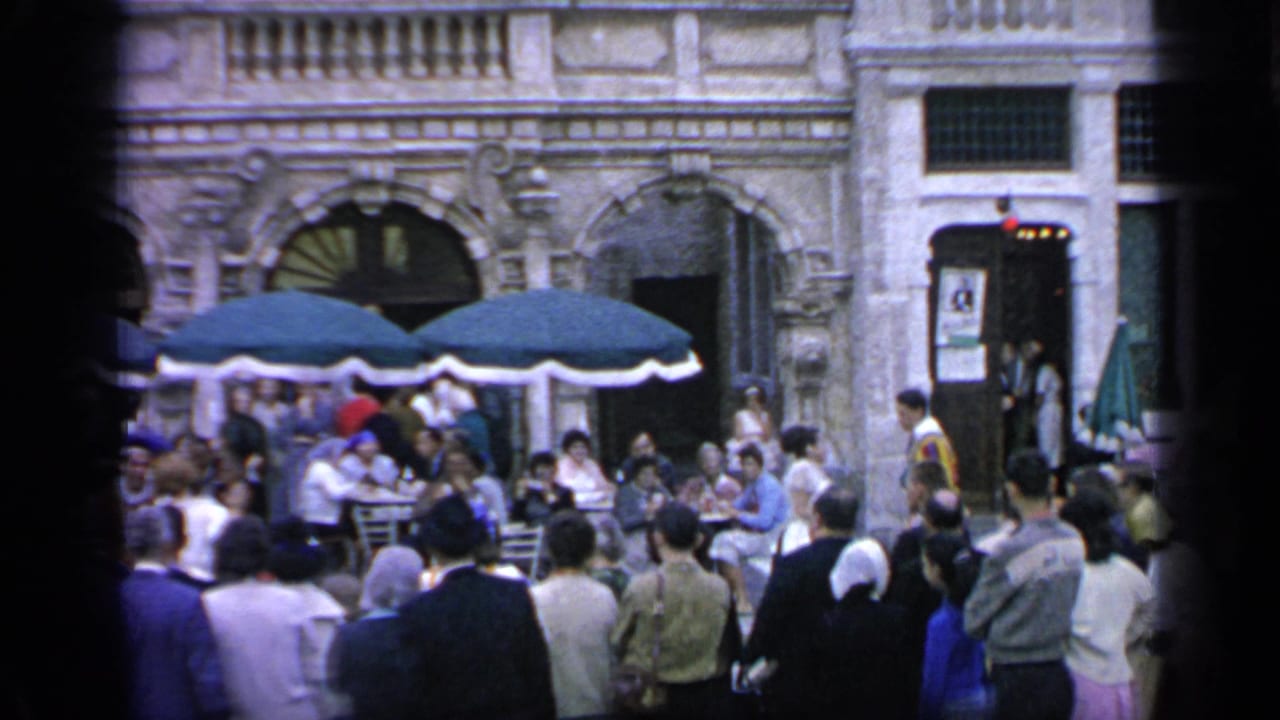 Crowd gathered around outdoor cafe tables under green umbrellas beside ornate arched facades.