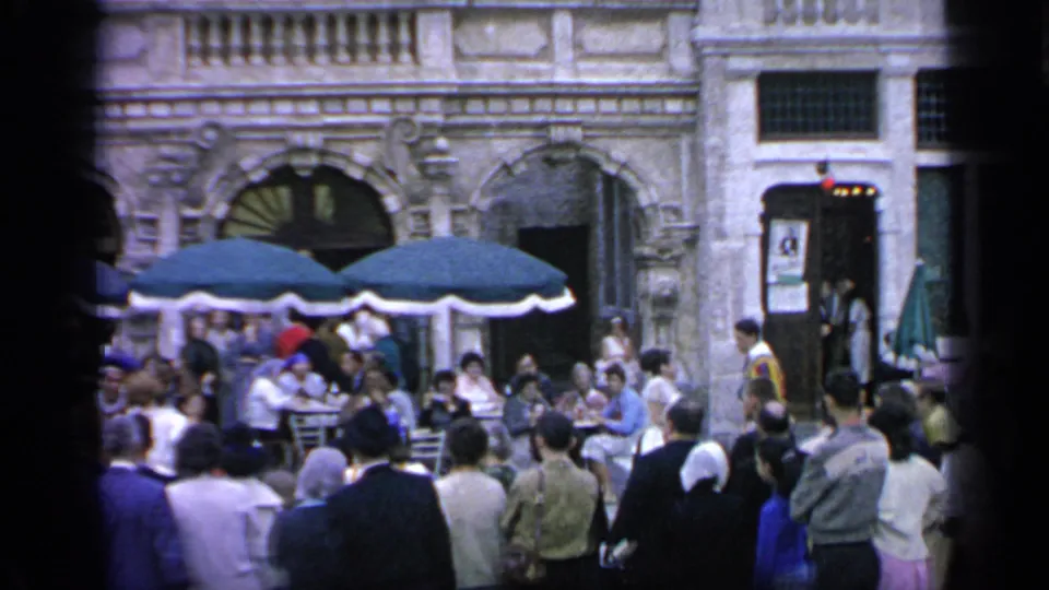 1964 Flushing Meadows home-movie frame showing visitors around outdoor tables with green umbrellas and decorative arched building fronts.