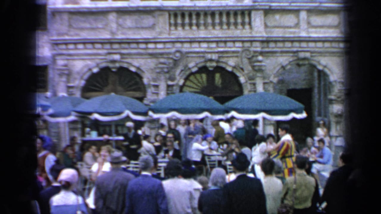 1964 Flushing Meadows footage frame with visitors dining and watching activity near green umbrellas and arched architectural details.