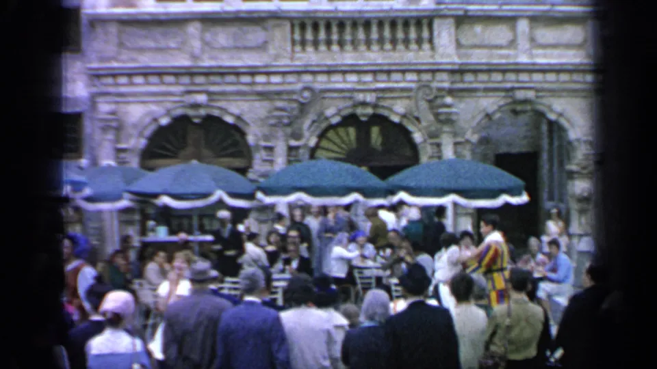 1964 Flushing Meadows footage frame with visitors dining and watching activity near green umbrellas and arched architectural details.
