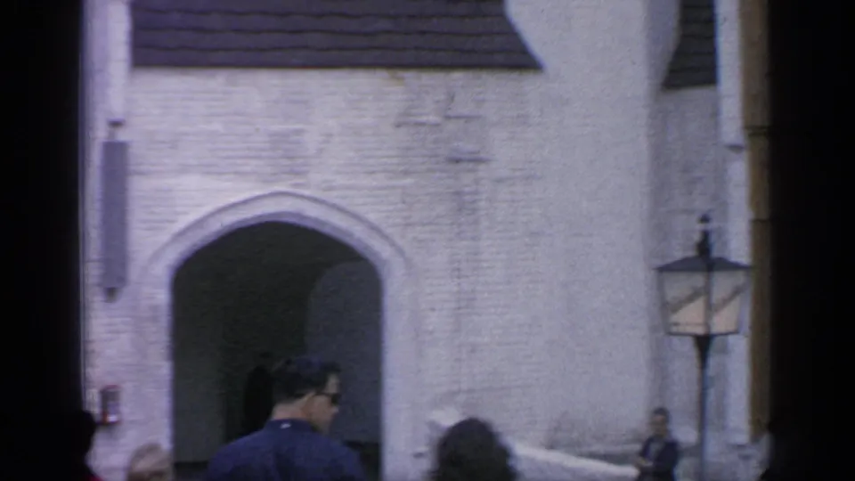 1964 Flushing Meadows home-movie frame showing a pale arched gateway, passing visitors, and a lantern on the wall.