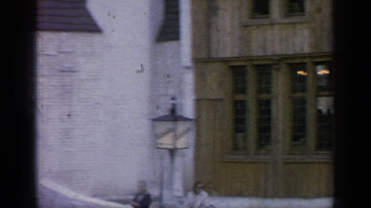 Two women near a lamppost and low wall in front of timber-framed windows.