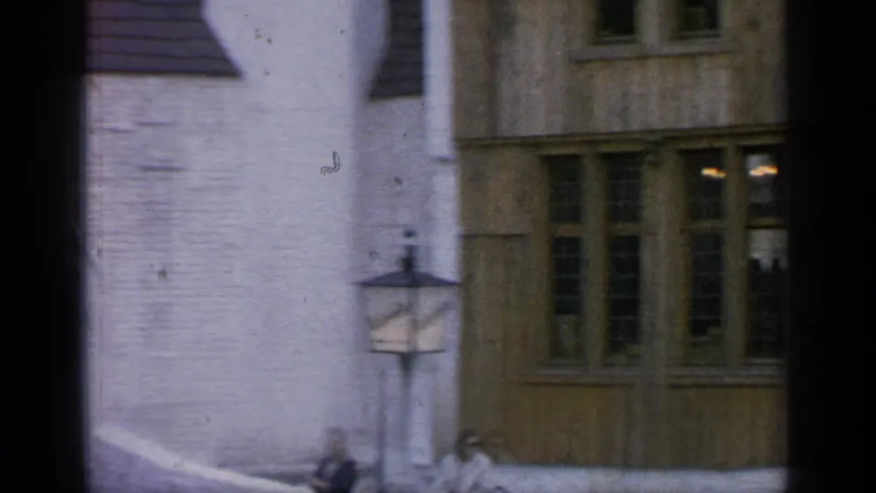 1964 Flushing Meadows home-movie frame showing two women by a lamppost and low wall outside white and timber-framed buildings.