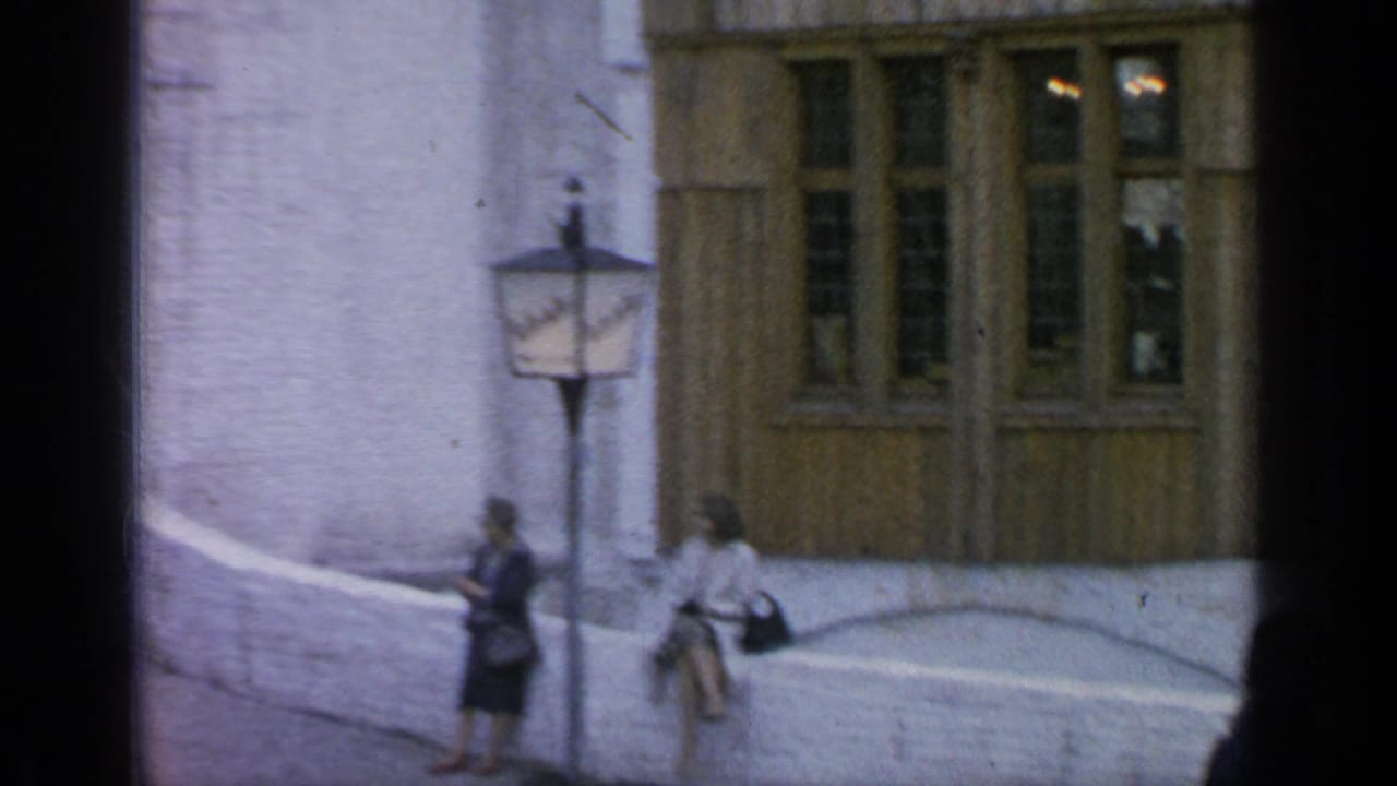 Two women with handbags stand or sit near a lamppost beside a pale wall.