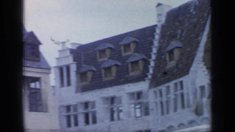 Home-movie view of fairground-style buildings with steep dark roofs, dormer windows, white walls, and partial film gate borders.