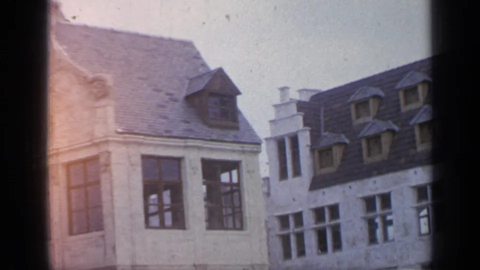 Flushing Meadows home-movie frame showing white facades, dark roofs, dormer windows, and a reddish exposure flare across the scanned 8mm image.