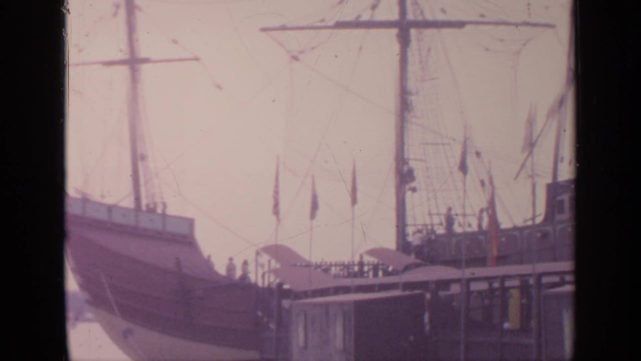 Tall-masted period-style ship at a dock with flags and rigging visible.